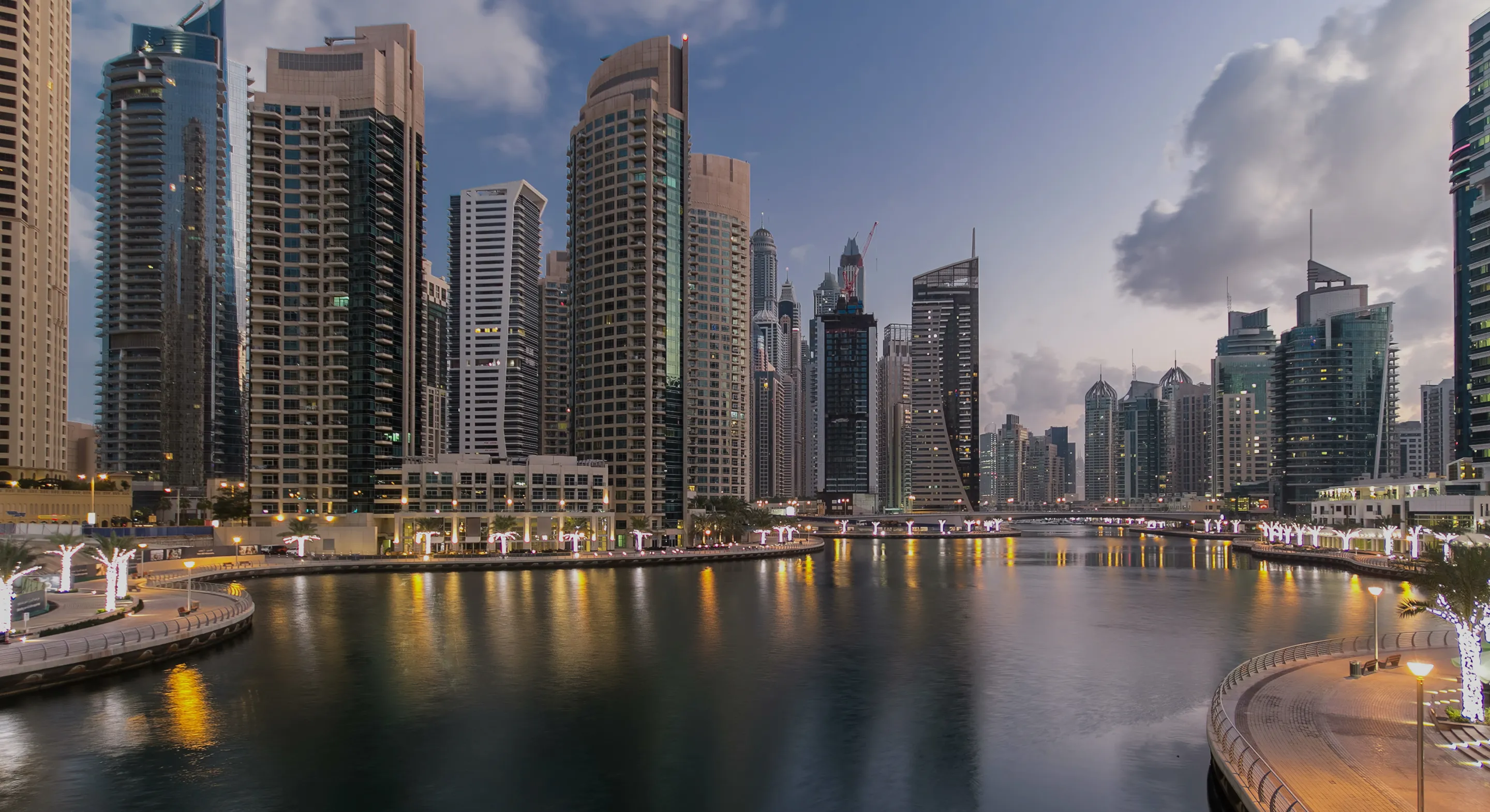 Evening view of a modern city skyline with skyscrapers reflecting in a calm water canal lined with lit palm trees and pedestrian walkways.