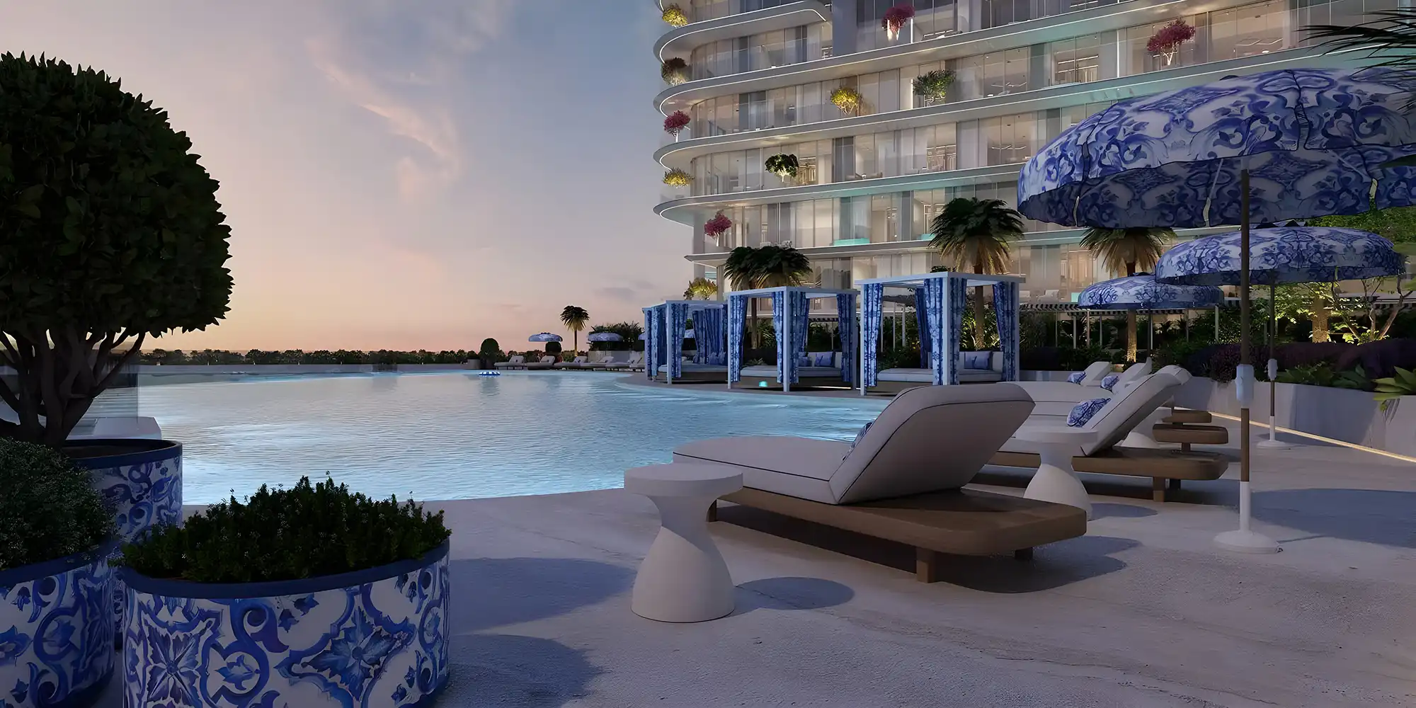 Modern poolside area at sunset with lounge chairs, blue patterned umbrellas, and cabanas beside a curved glass building.
