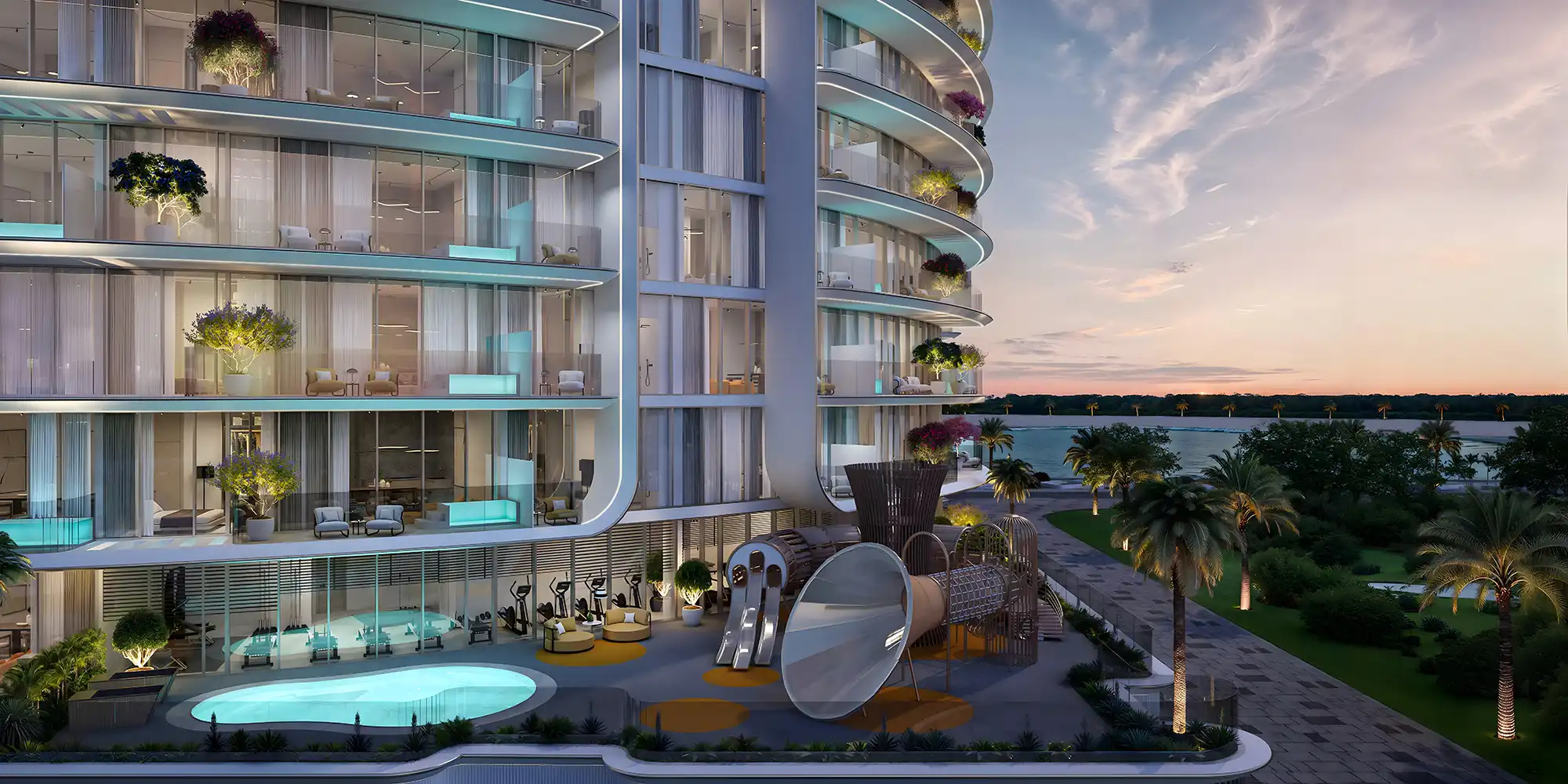 Modern curved building with illuminated balconies overlooking a pool, playground, palm trees, and a lake at sunset.
