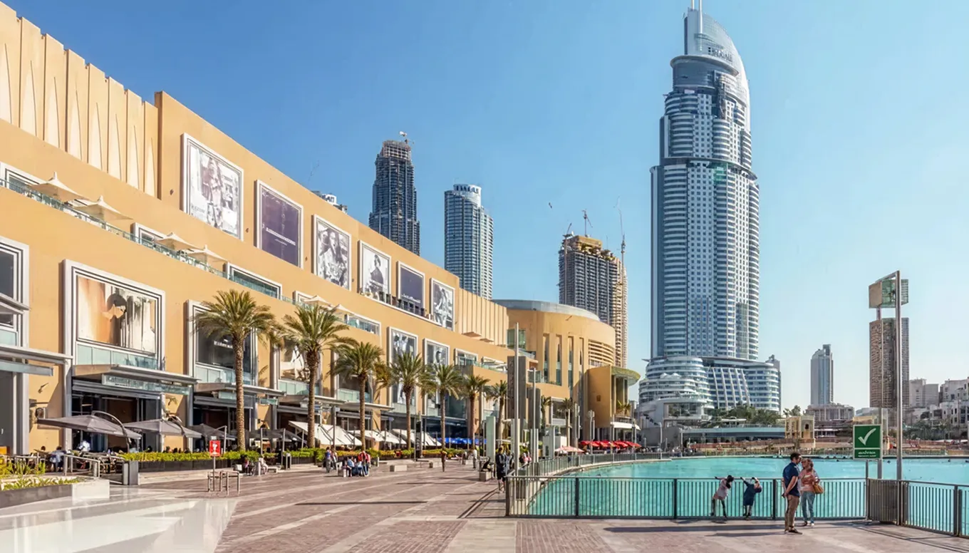 Waterfront promenade with palm trees, outdoor seating, and modern skyscrapers under a clear blue sky.