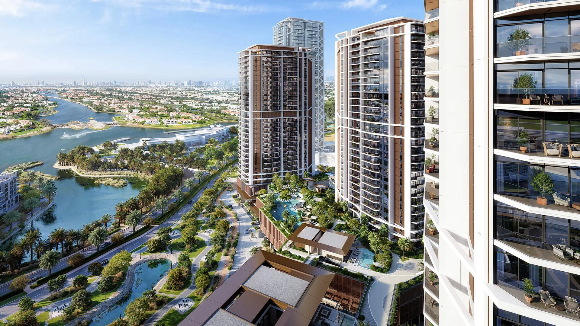 Modern high-rise residential buildings with glass balconies overlooking a landscaped area with pools and palm trees beside a large river.