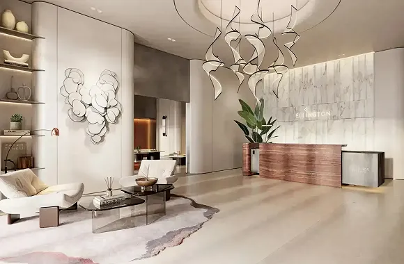 Modern hotel lobby with sculptural wall art, two white lounge chairs, a glass coffee table, a wavy beige and grey rug, and a wooden reception desk with green plants.