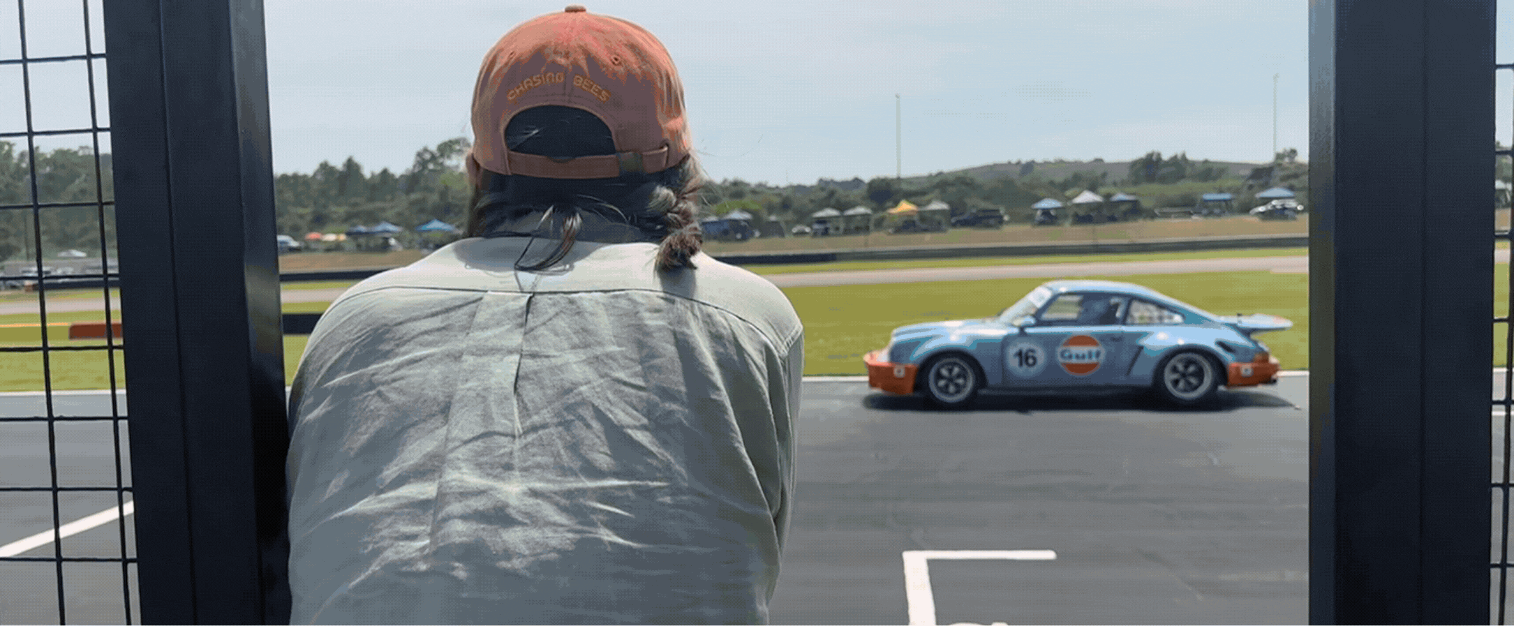 Person wearing a red cap and light-colored shirt watching a vintage blue and orange race car with number 16 on a racetrack.