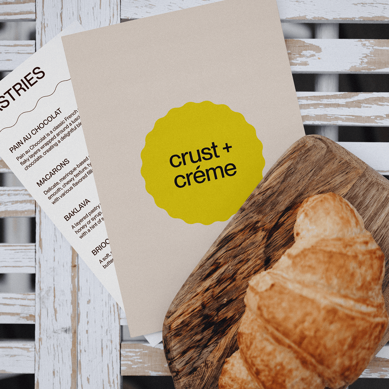 Croissant on a wooden board next to a menu card with a yellow logo reading 'crust + crème' on a white rustic table.