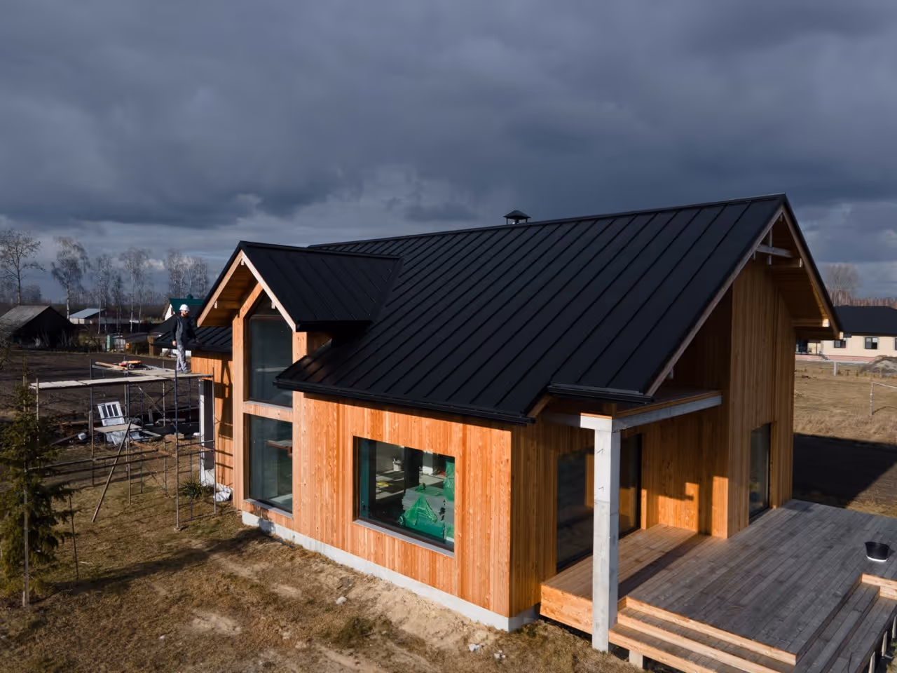 Wooden house with black metal roof under a cloudy sky, with a worker standing on scaffolding on the side.