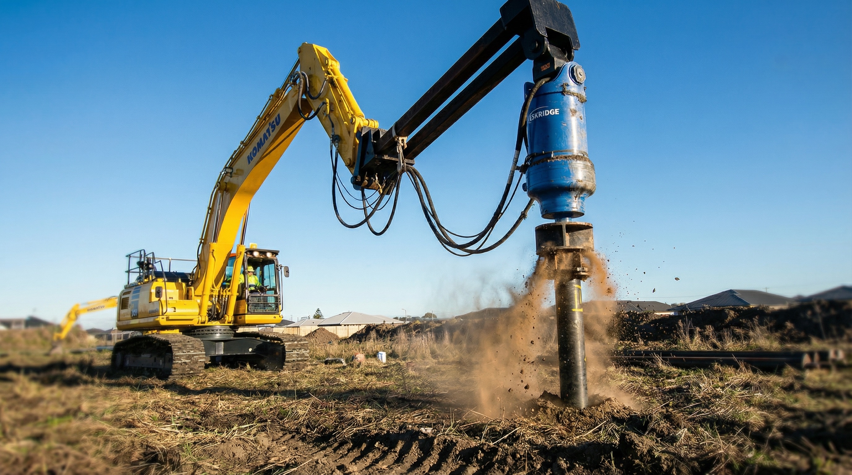 Yellow Komatsu excavator with a blue drilling attachment active on a construction site on a clear day.