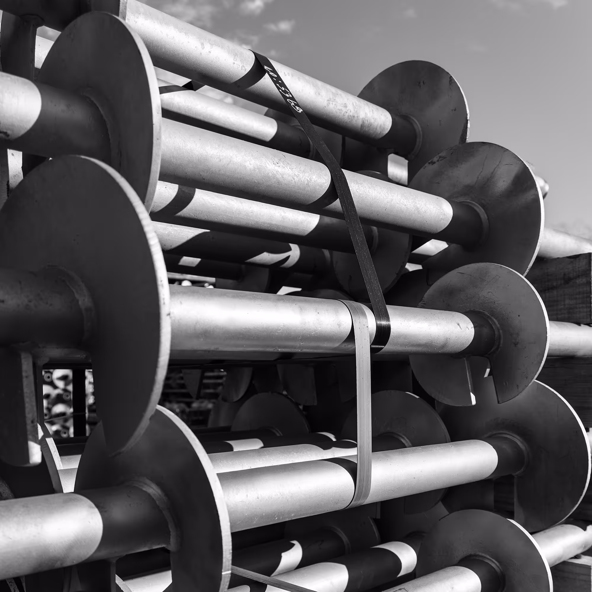 Stacked metal screw piles with wide circular plates, secured with straps under a partly cloudy sky.