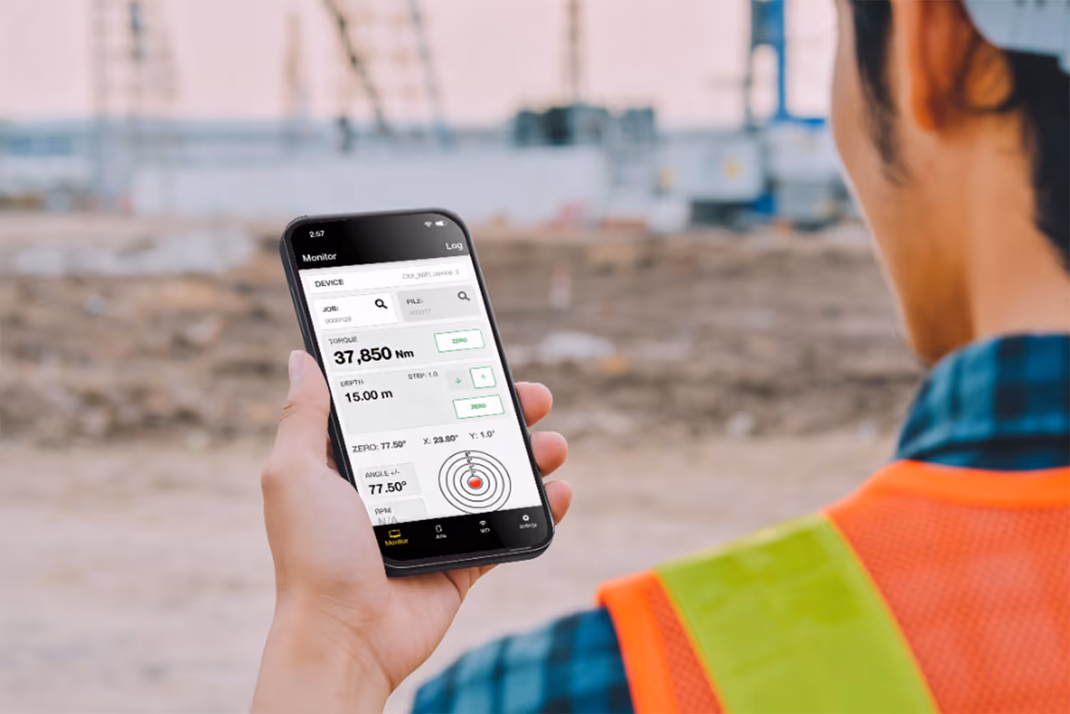 Person wearing an orange safety vest holding a smartphone displaying a torque and depth monitoring app on a construction site.