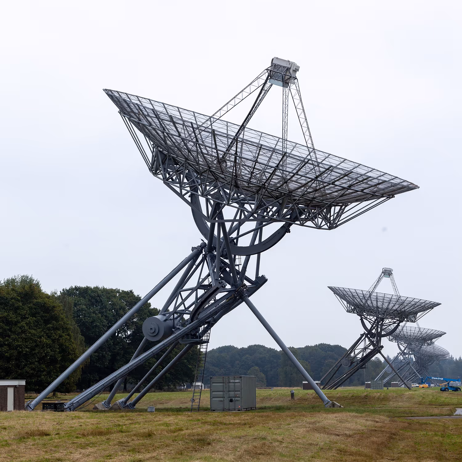 A row of large metal satellite dishes with mesh parabolic reflectors in an open grassy field under an overcast sky.