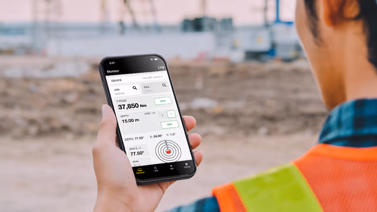 Person wearing an orange safety vest holding a smartphone displaying a torque and depth monitoring app on a construction site.