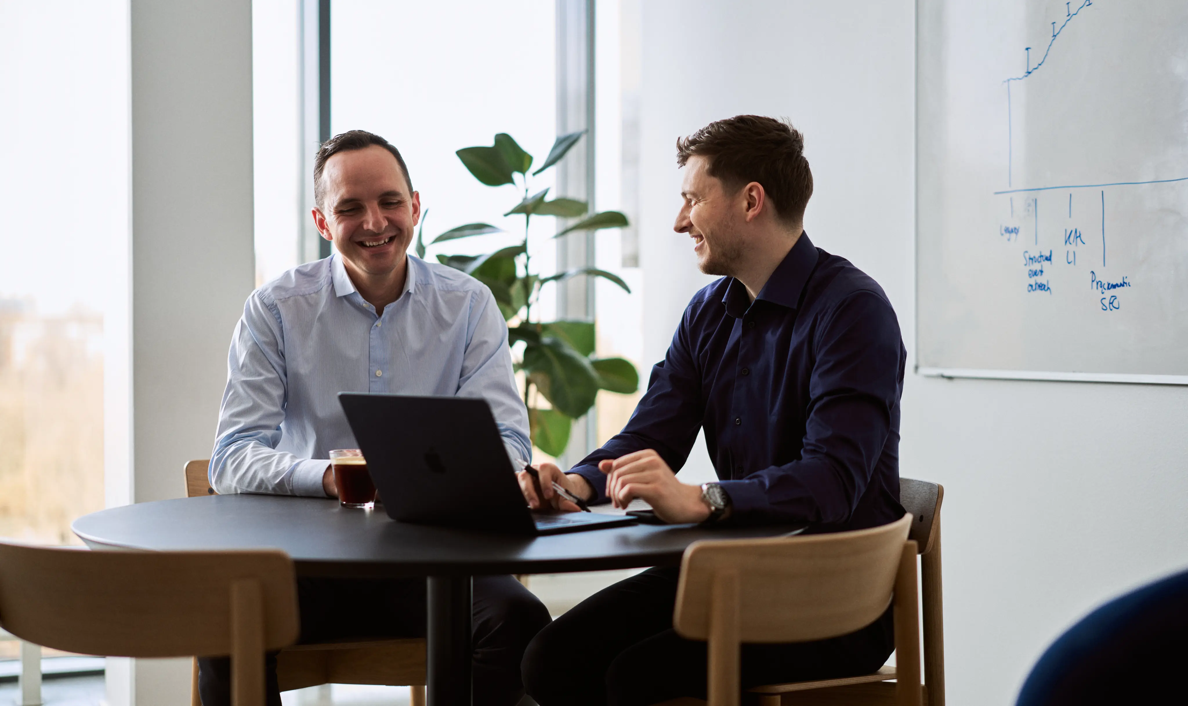 Two men smiling and talking while sitting at a round table with a laptop in a bright office room.