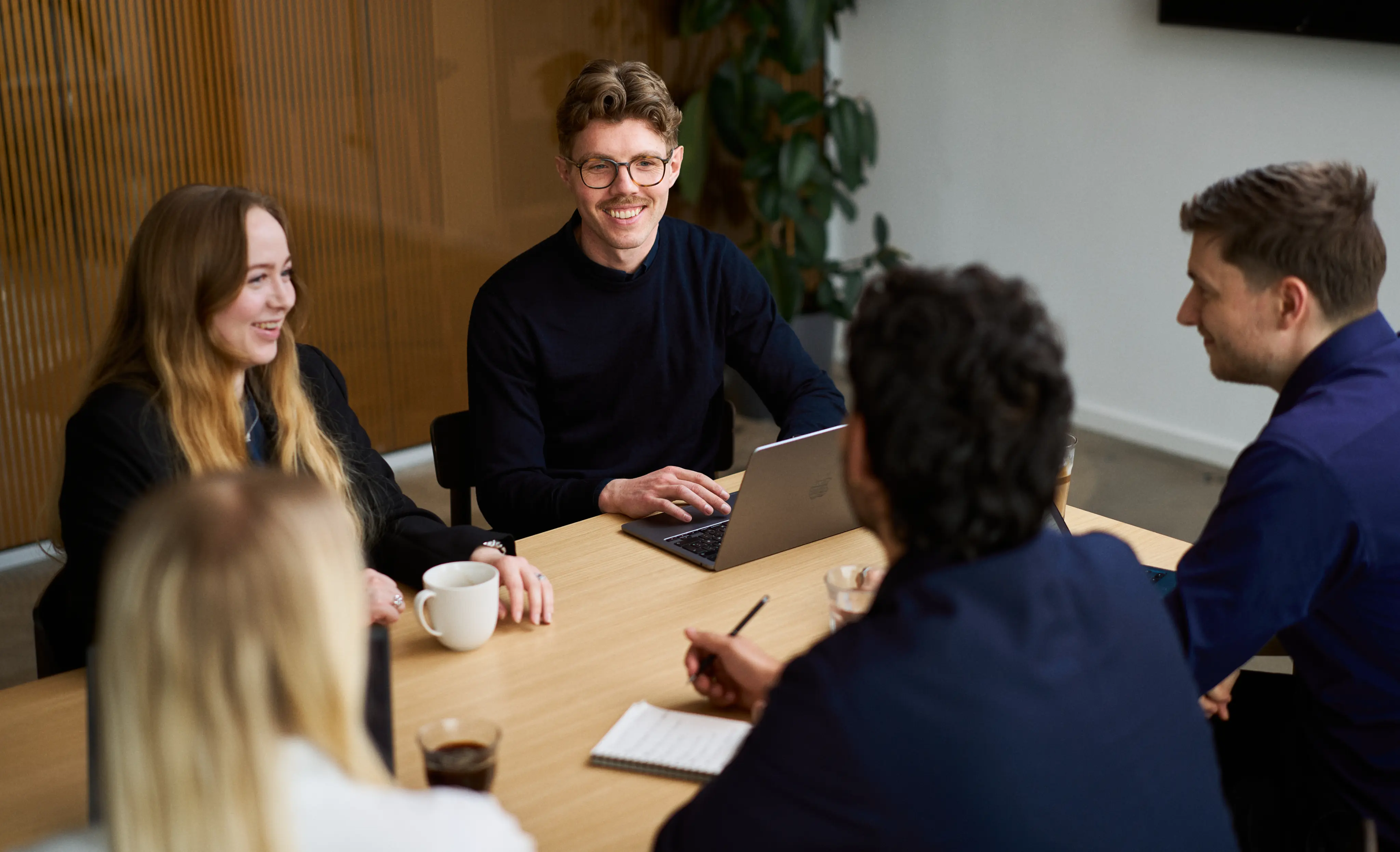 Three young professionals engaged in a meeting in a bright office with large windows, one woman standing and two seated at a wooden table with a laptop.