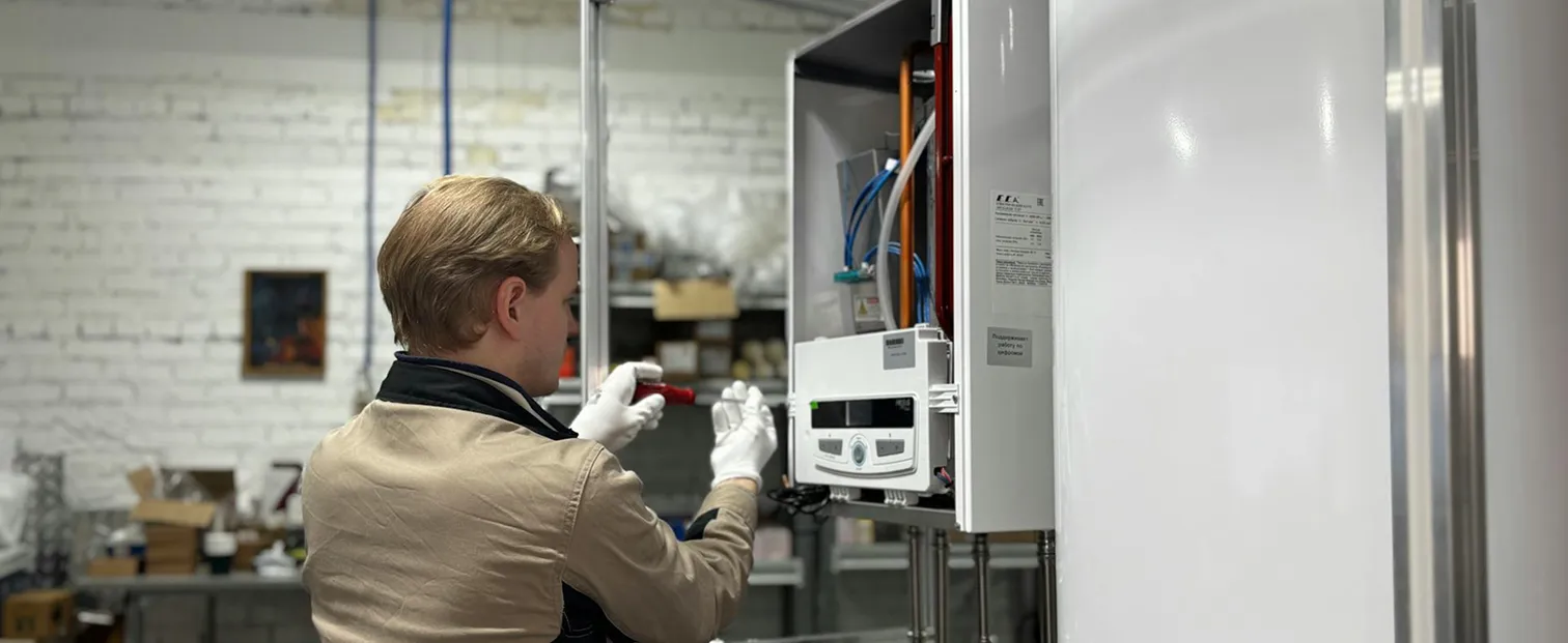 Technician wearing white gloves using a screwdriver to repair or maintain an open wall-mounted boiler unit in a workshop.