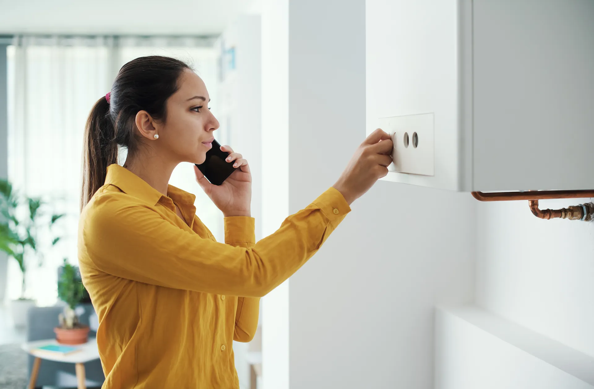 Woman in a yellow shirt adjusting a thermostat while talking on a smartphone in a modern home.