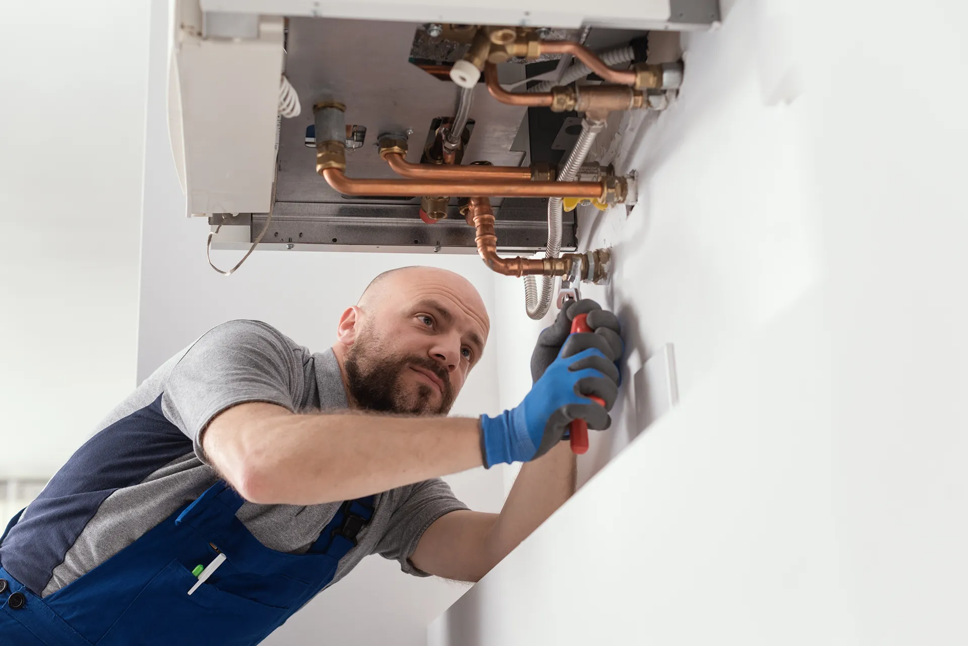 Technician in blue overalls and gloves repairing copper pipes under a boiler mounted on a white wall.