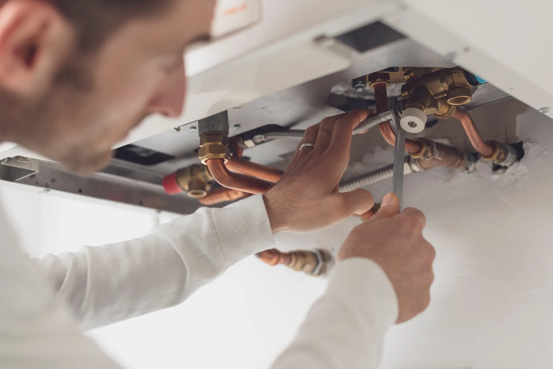 Man using a wrench to adjust copper pipes under a boiler or heating unit.