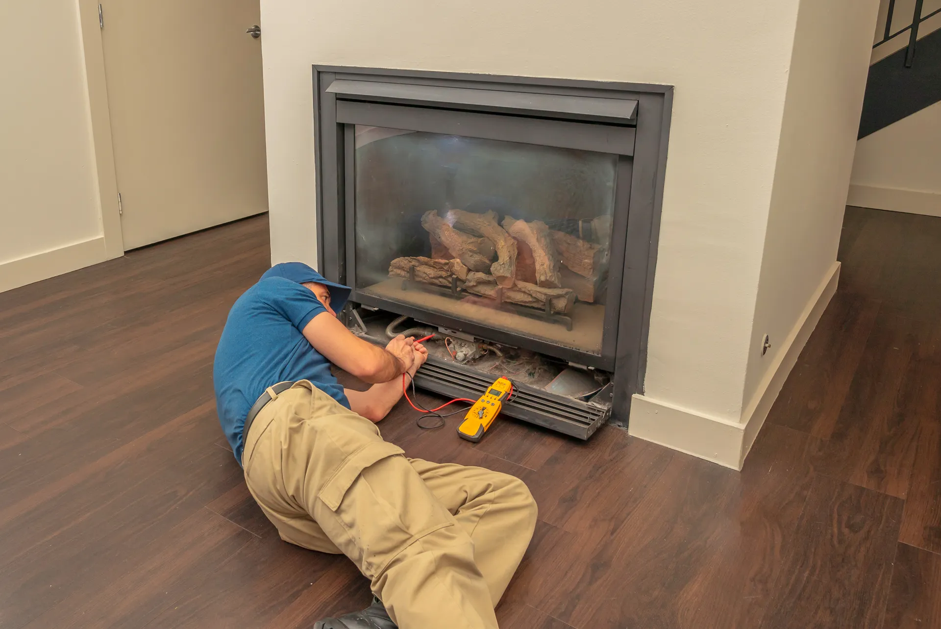Technician lying on the floor repairing a gas fireplace using a multimeter.