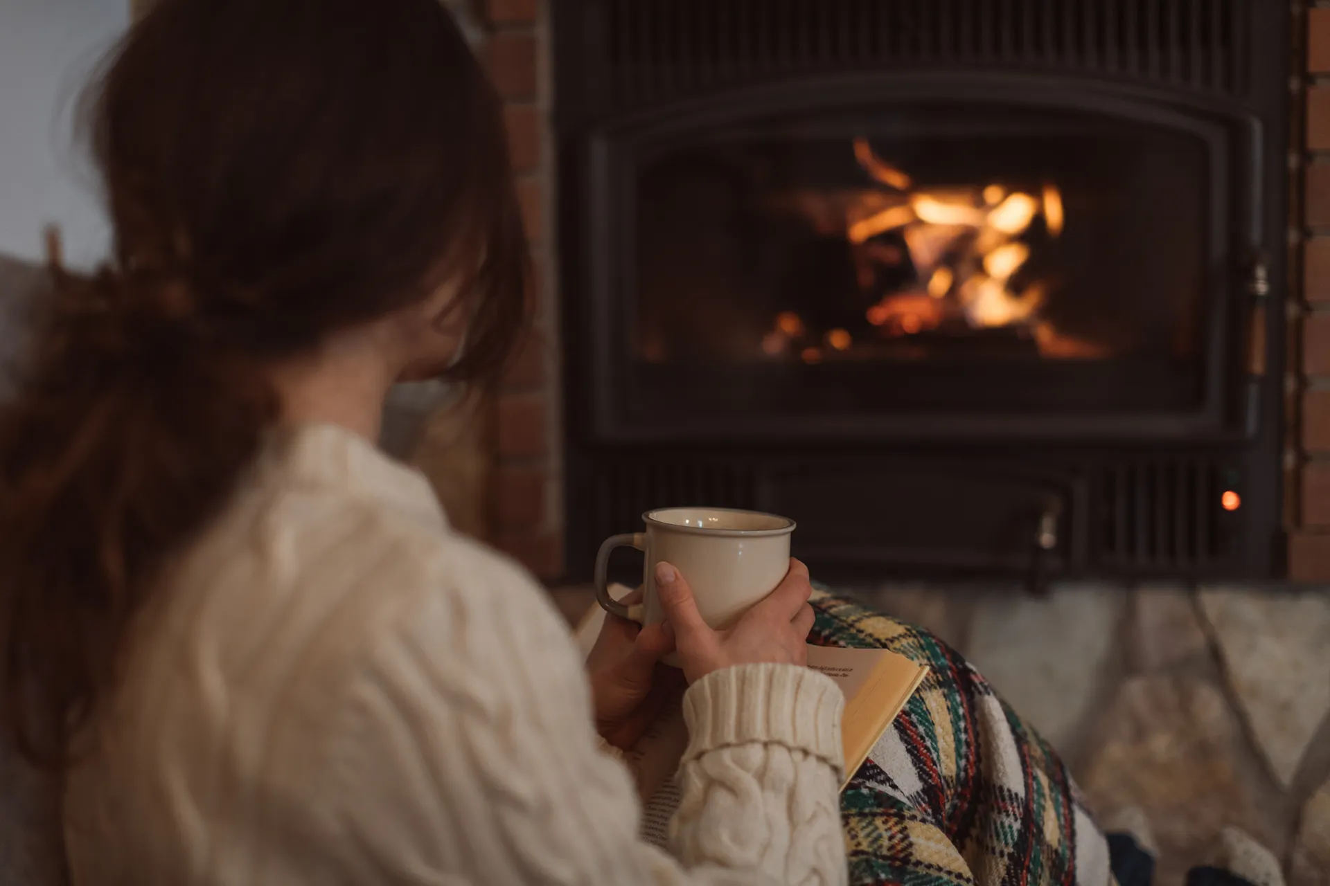 Person in a cozy sweater holding a mug and sitting with a book near a lit fireplace.