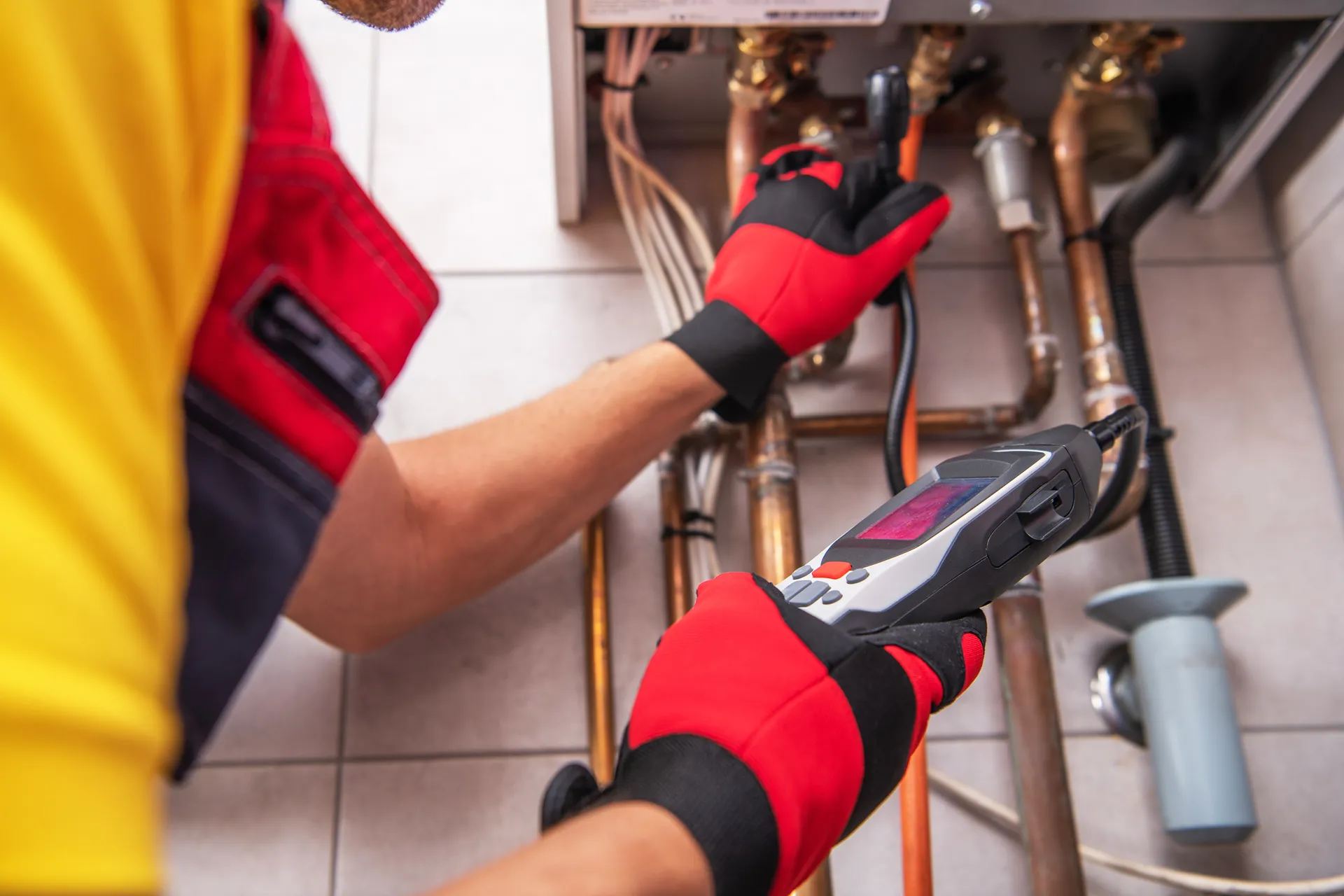 Technician wearing red and black gloves using a diagnostic device to inspect copper pipes of a gas boiler system.
