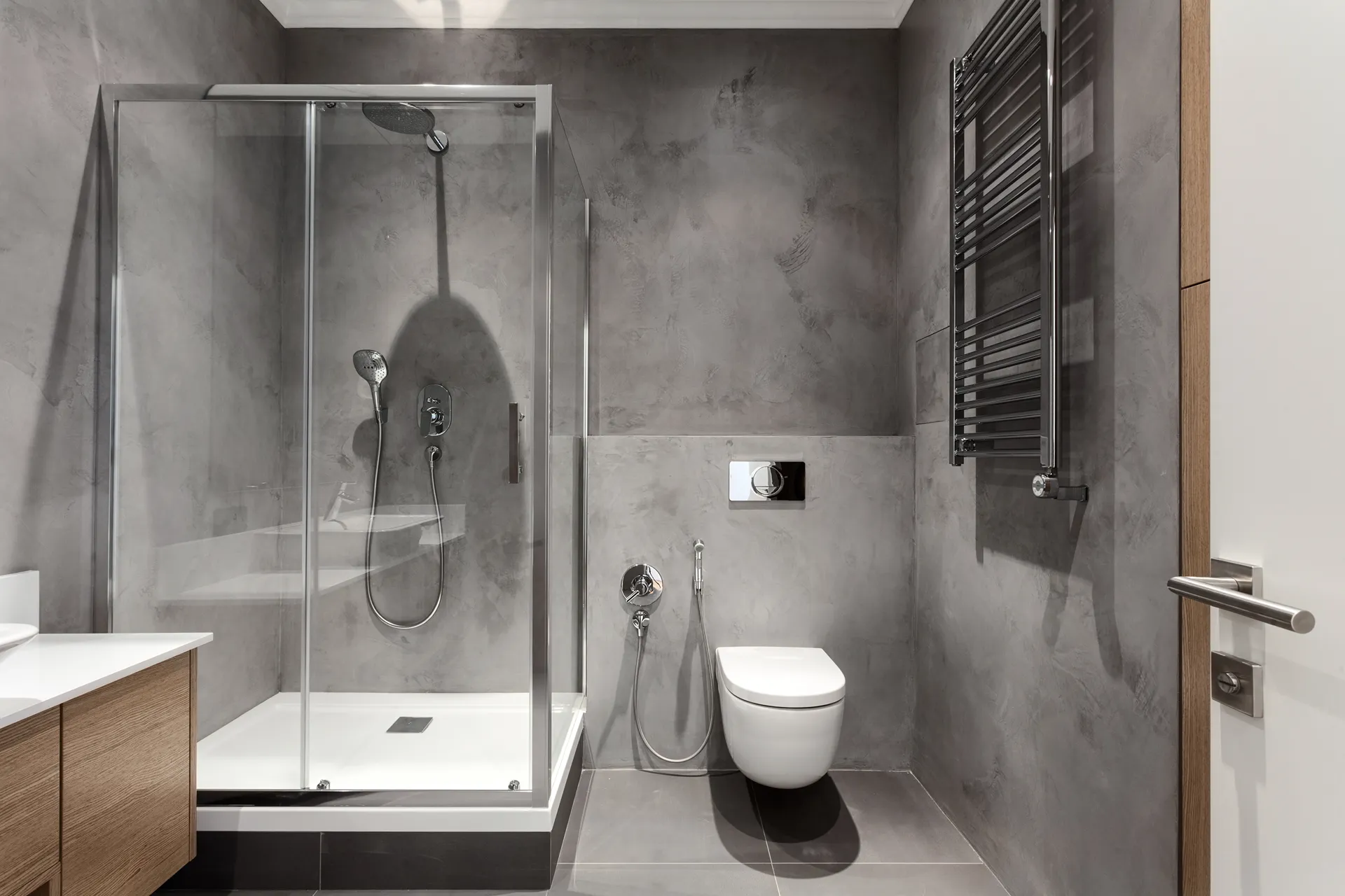Modern bathroom with grey concrete walls, glass-enclosed shower, wall-mounted toilet, wooden vanity, and chrome towel radiator.