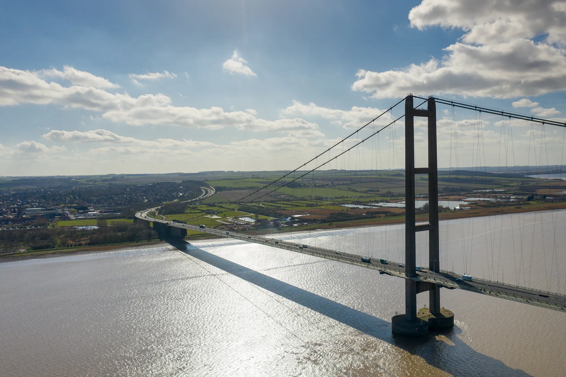 Aerial view of a long suspension bridge crossing a wide river with green fields and countryside in the background under a partly cloudy sky.