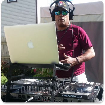 DJ wearing sunglasses, headphones, and a maroon shirt standing behind a Denon DJ mixer and a MacBook laptop outdoors.