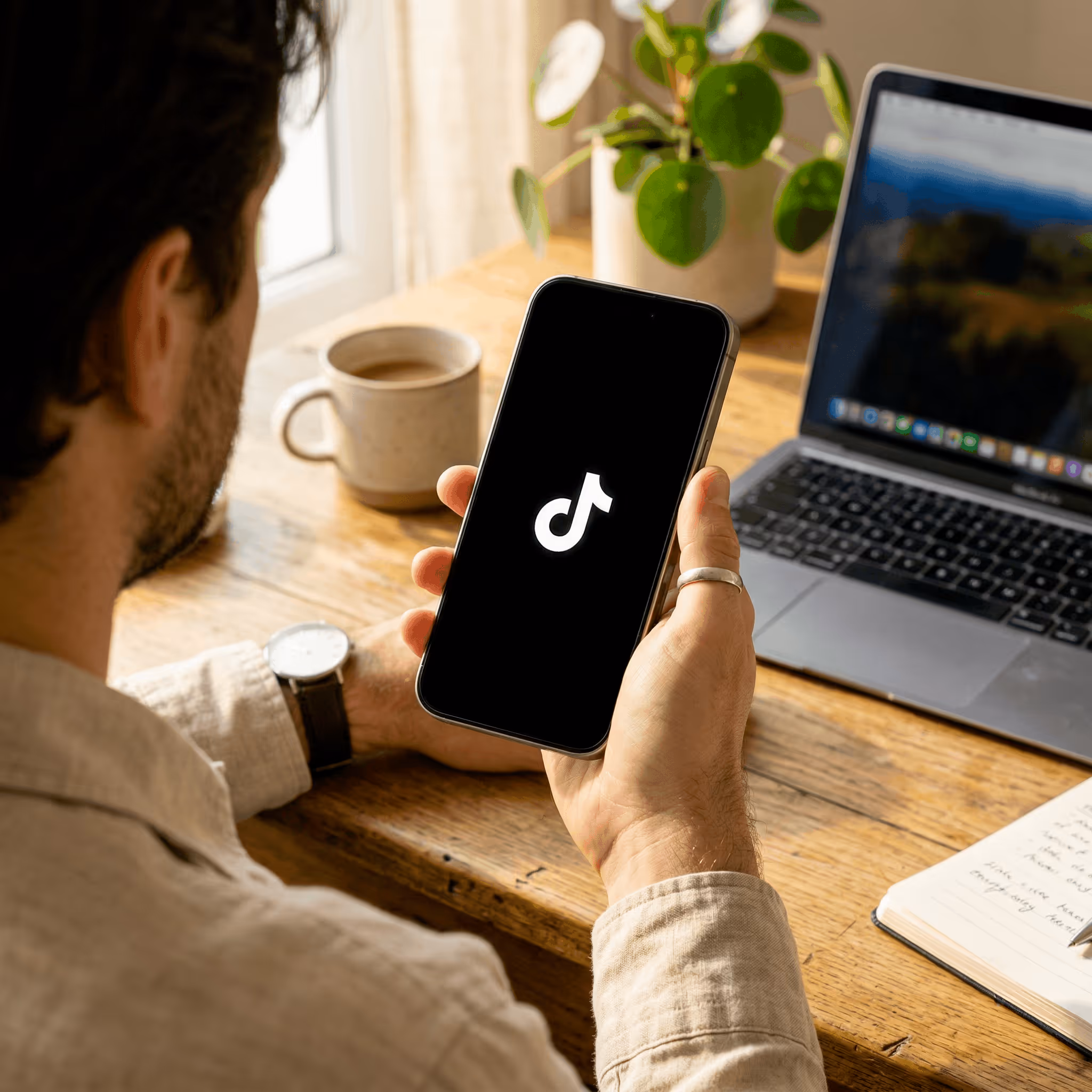 Un homme tenant un smartphone affichant le logo TikTok, assis à une table avec un ordinateur portable, une tasse de café et un carnet.