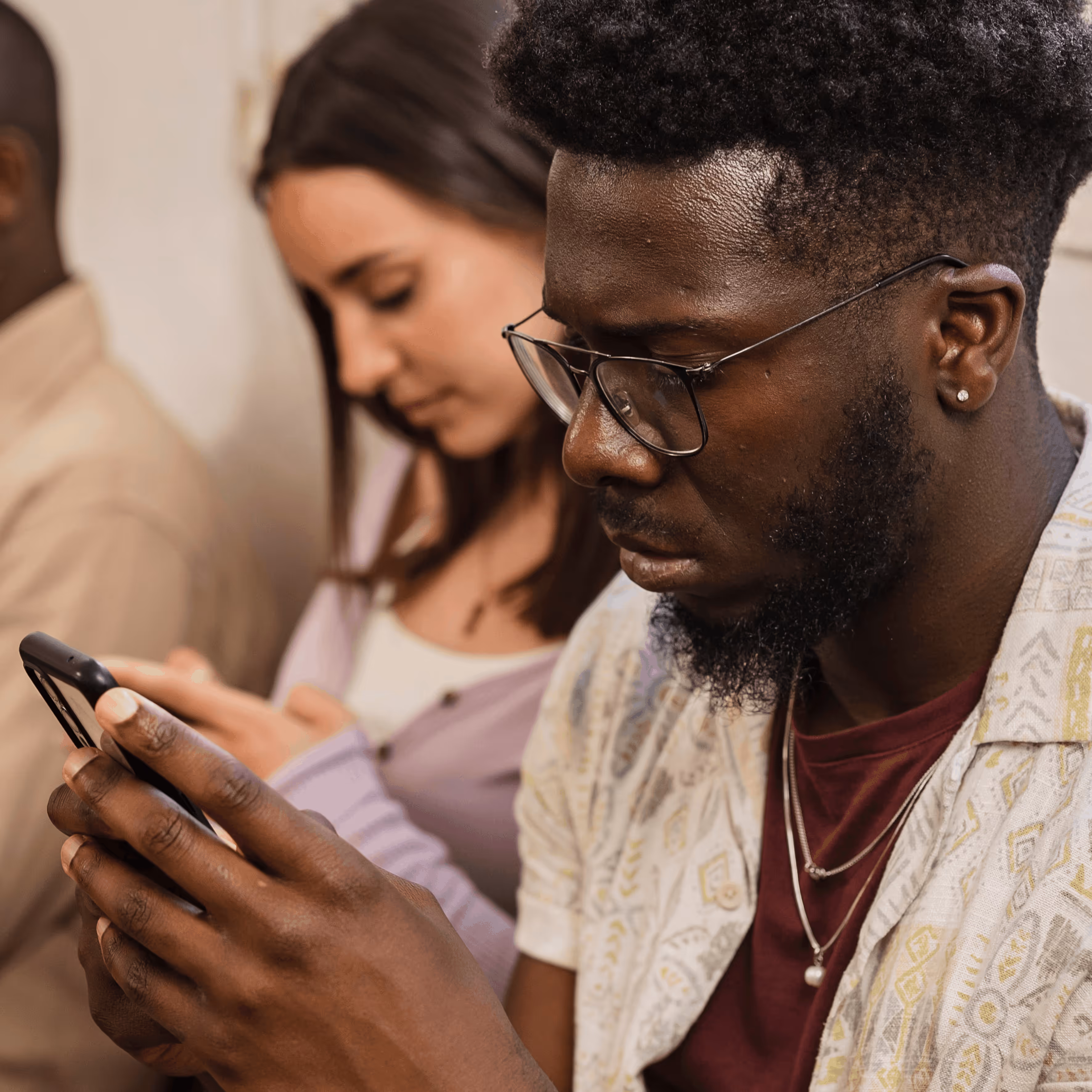 Jeune homme concentré portant des lunettes et regardant un téléphone portable, avec une femme floue en arrière-plan utilisant aussi un téléphone.