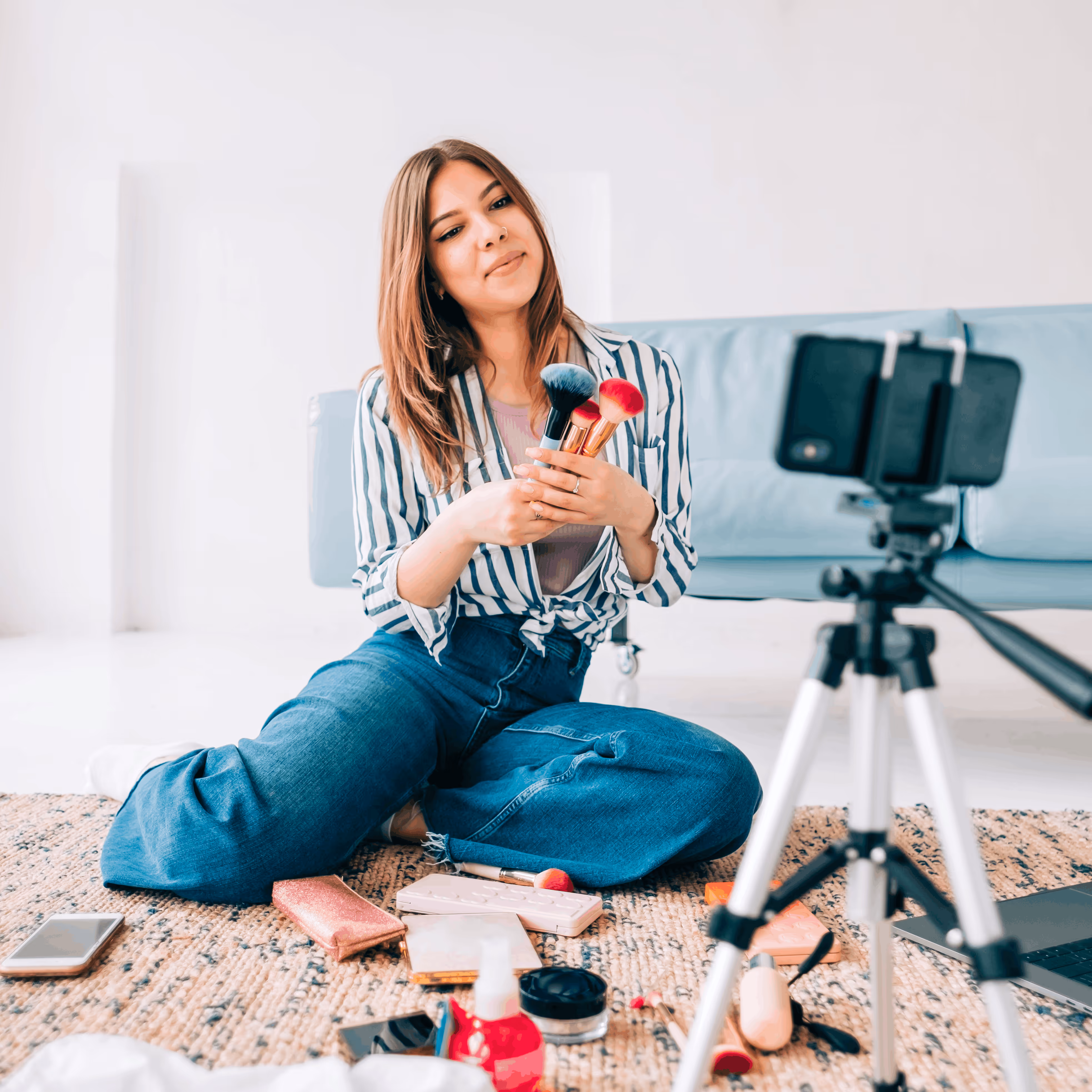 Une femme assise par terre tenant des pinceaux de maquillage, face à un smartphone sur un trépied, entourée de produits cosmétiques.