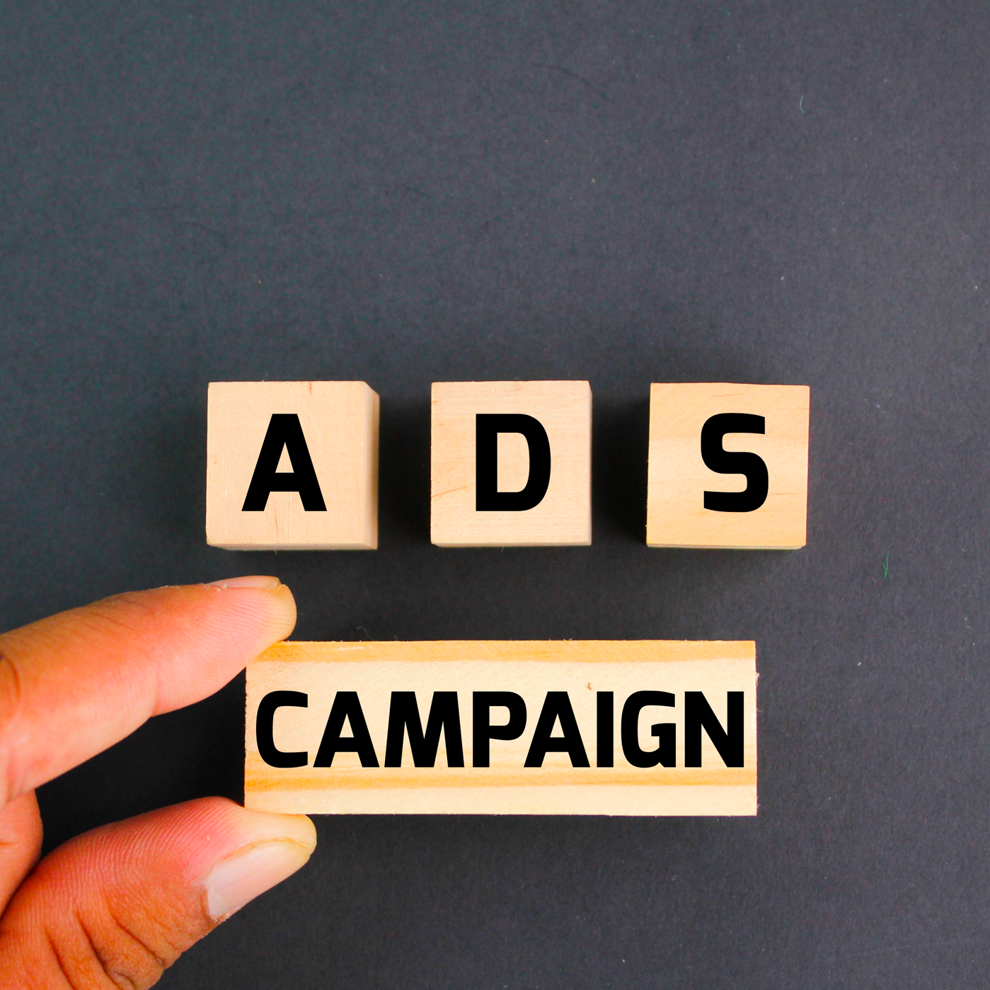 Un doigt tenant un bloc en bois avec le mot CAMPAGNE sous trois blocs avec les lettres ADS sur fond noir.