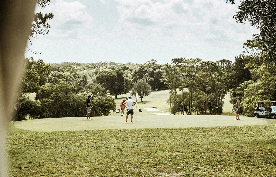 Four golfers on a putting green surrounded by trees under a partly cloudy sky.