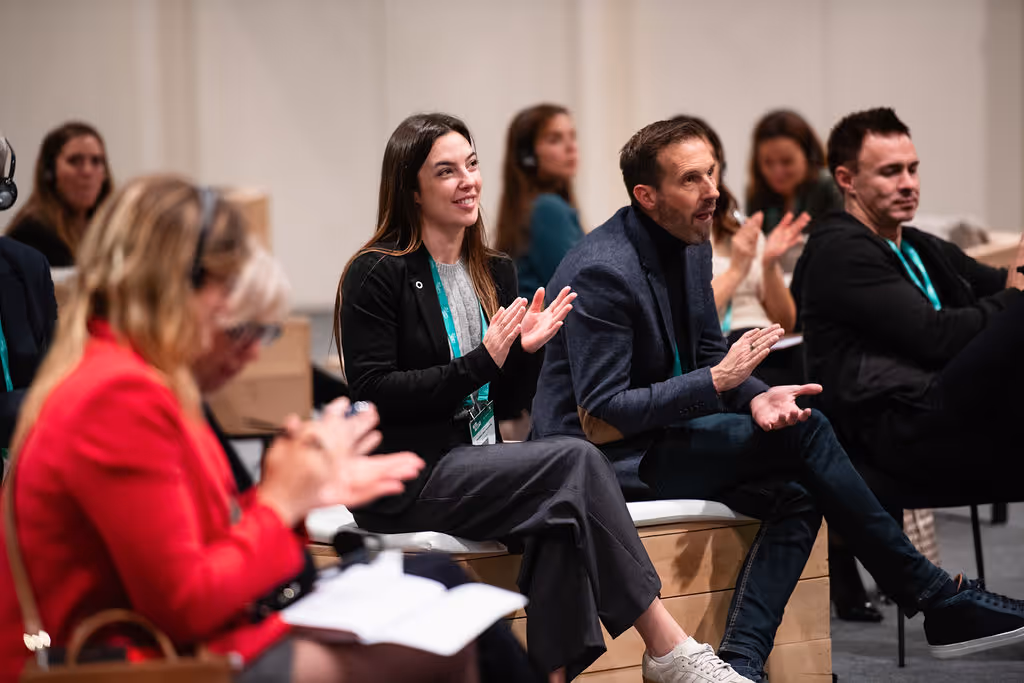People seated indoors, attentively listening and clapping during a presentation or event.