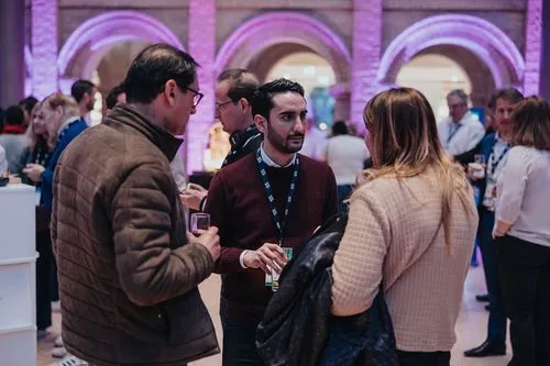 Three people engage in conversation at a social event, surrounded by others in a well-lit room.