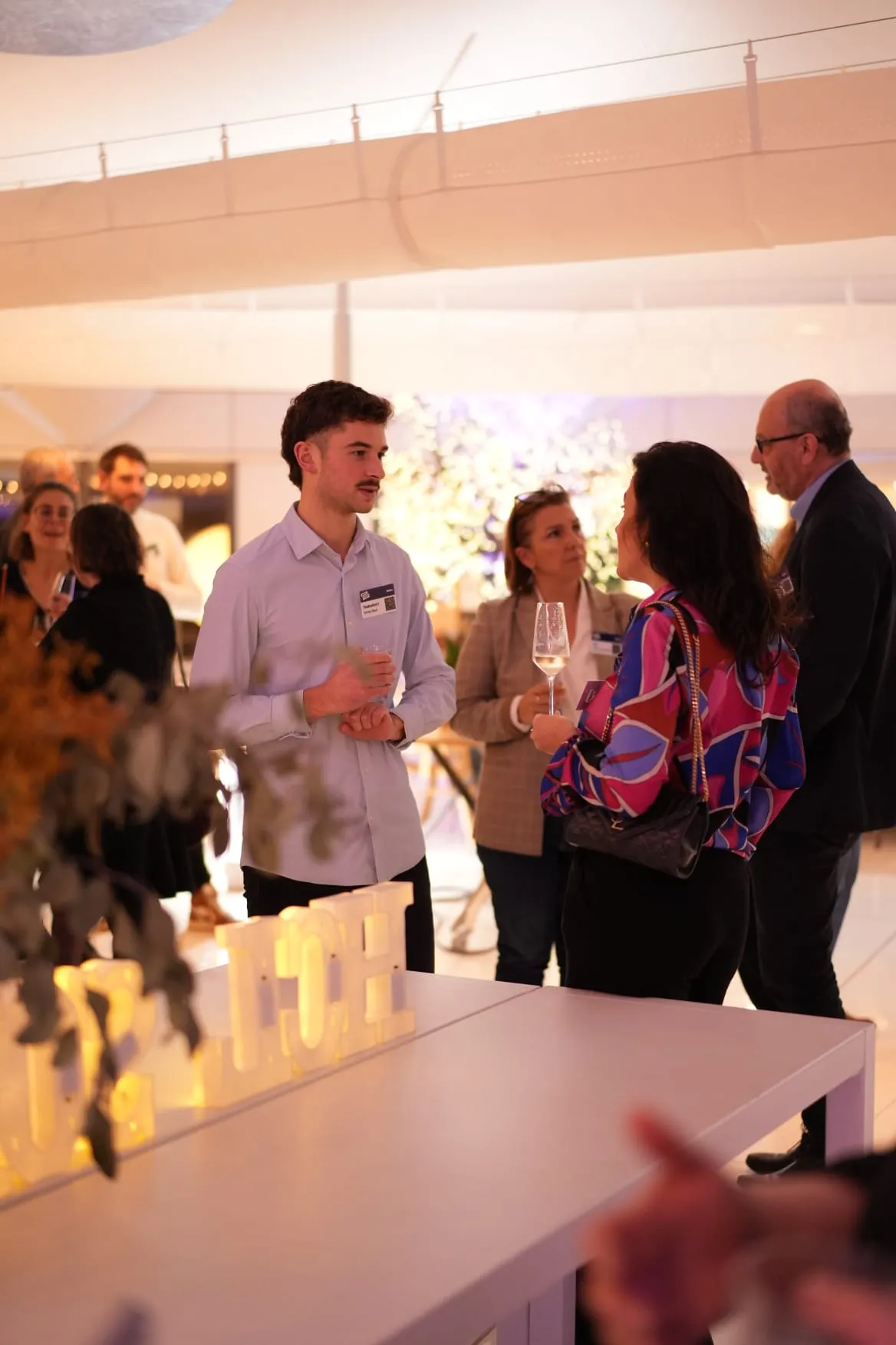 A man and a woman are talking at an indoor social gathering, holding glasses.
