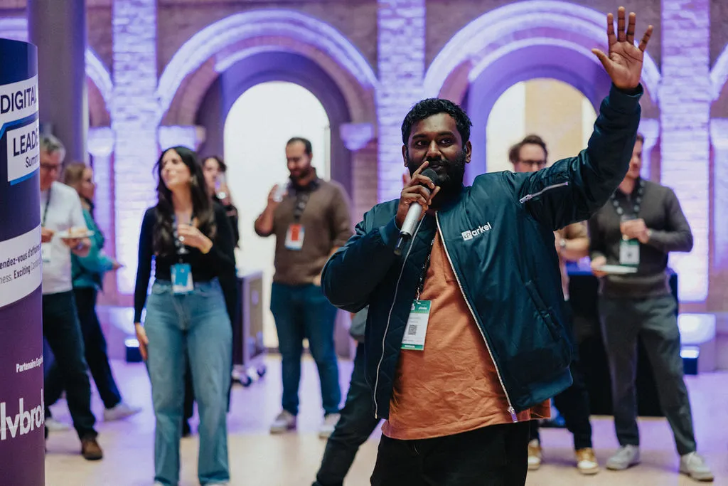 Man speaking into a microphone, raising his hand, while people listen in a large room.