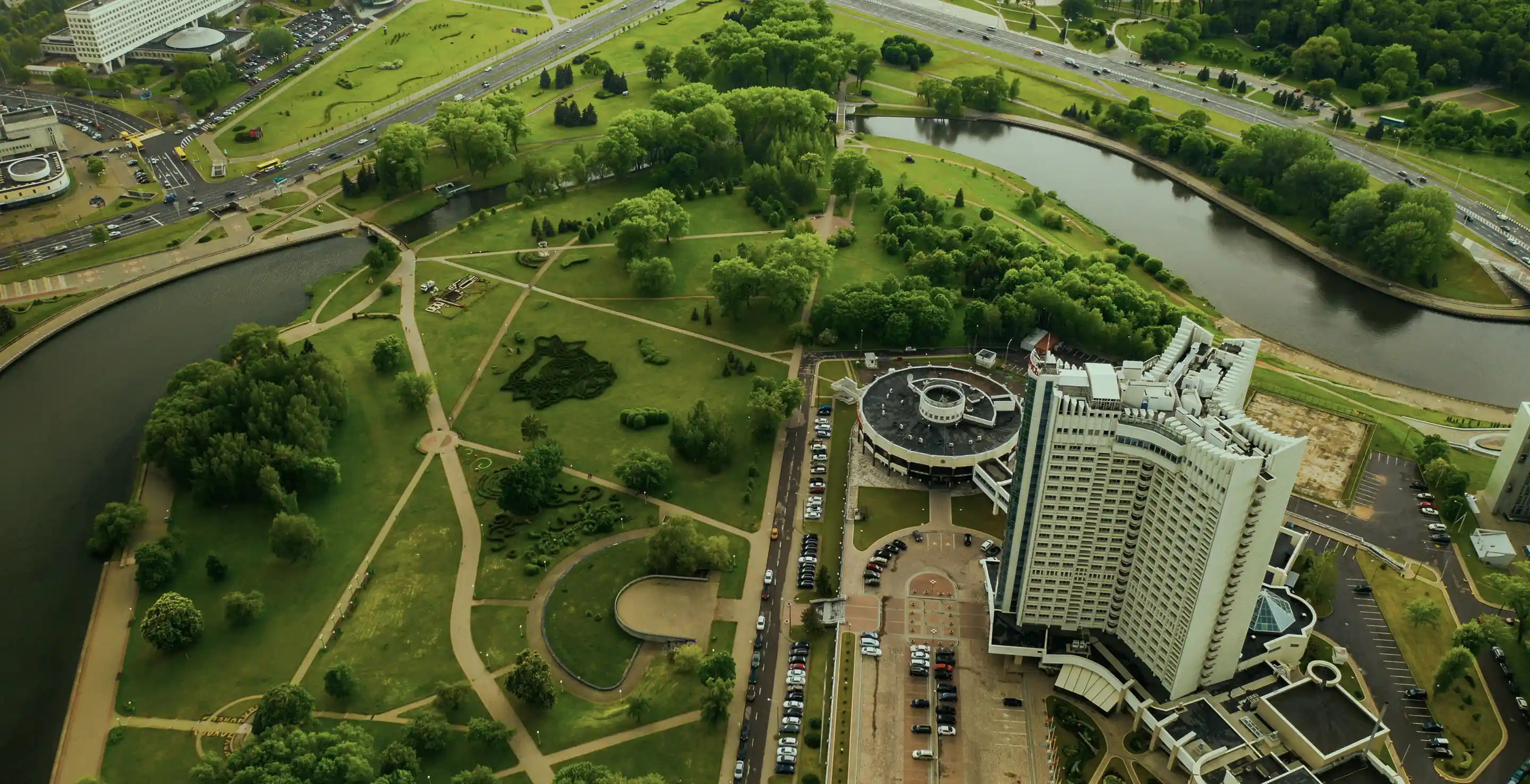 Veduta aerea di un grande edificio moderno accanto a un parco verde con sentieri e un fiume sinuoso.