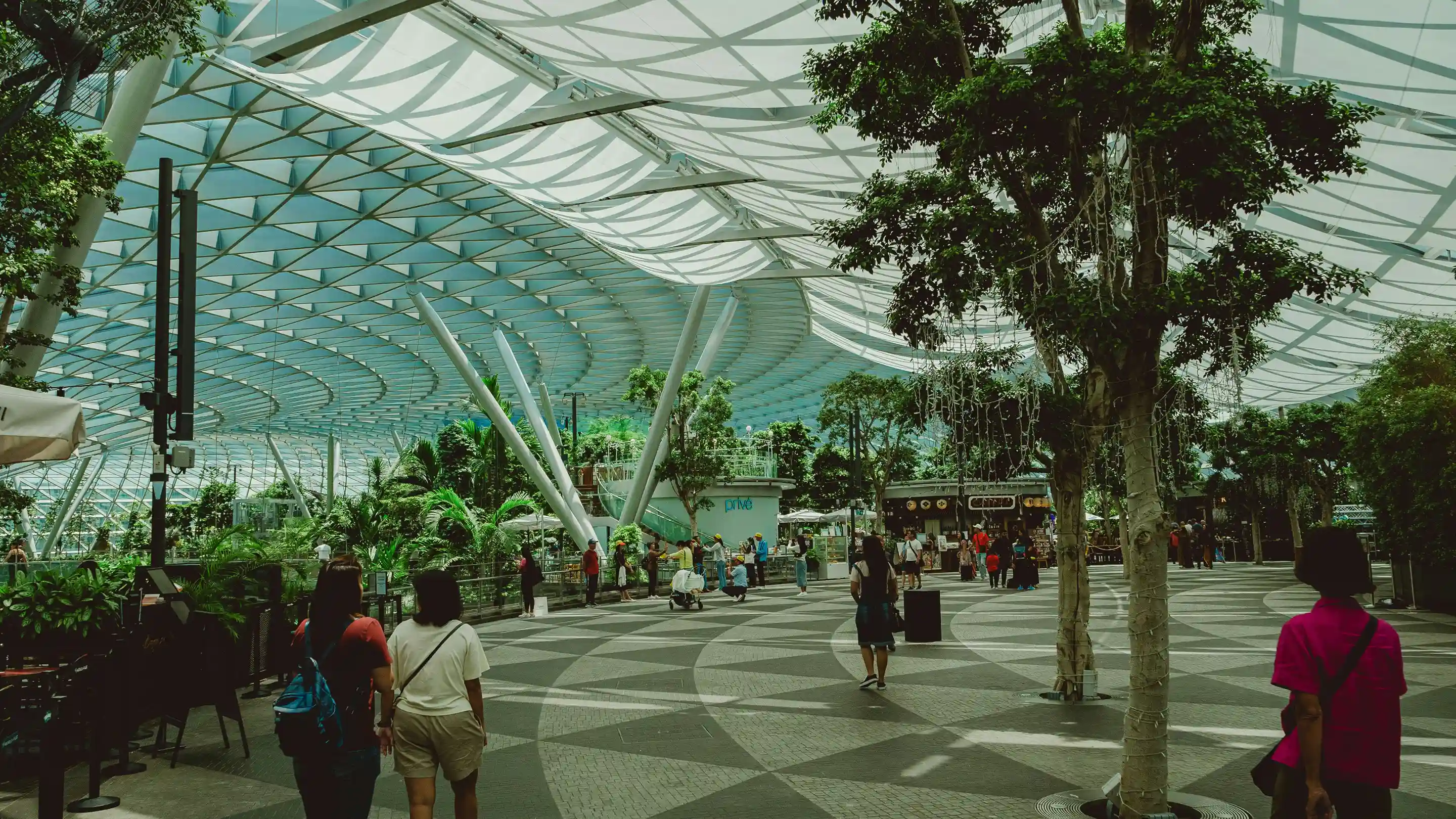 Area interna di un grande centro commerciale con ampio soffitto in vetro e metallo, piante verdi e persone che camminano.