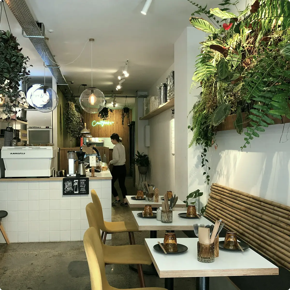Interno di un caffè moderno con pareti bianche, piante verdi appese, sedie gialle e tavoli apparecchiati con bicchieri e posate.
