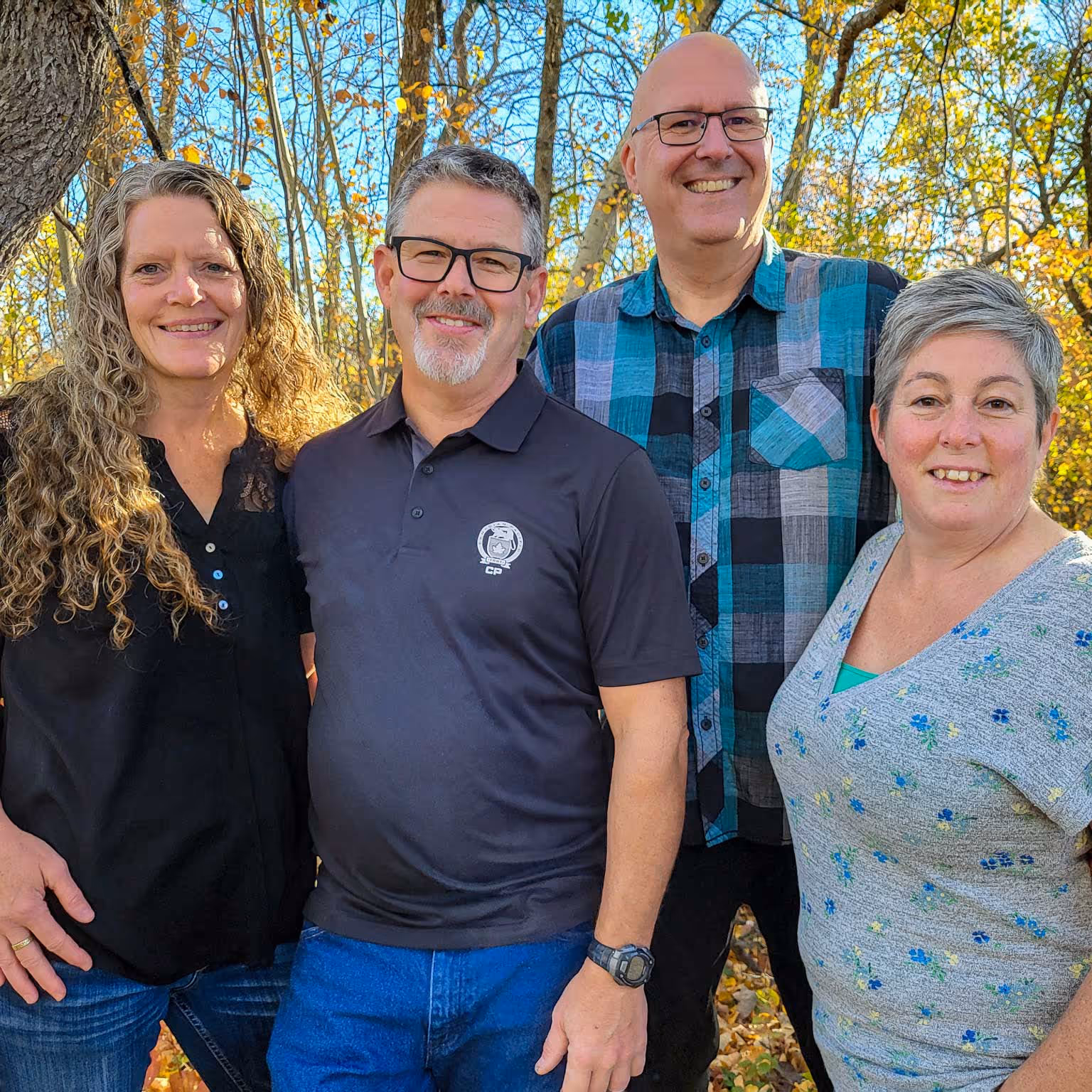 Mike and Tracy Cole, Curtis and Teresa Benson, owners of Colben Enterprises, smiling outdoors with autumn trees and blue sky in the background.