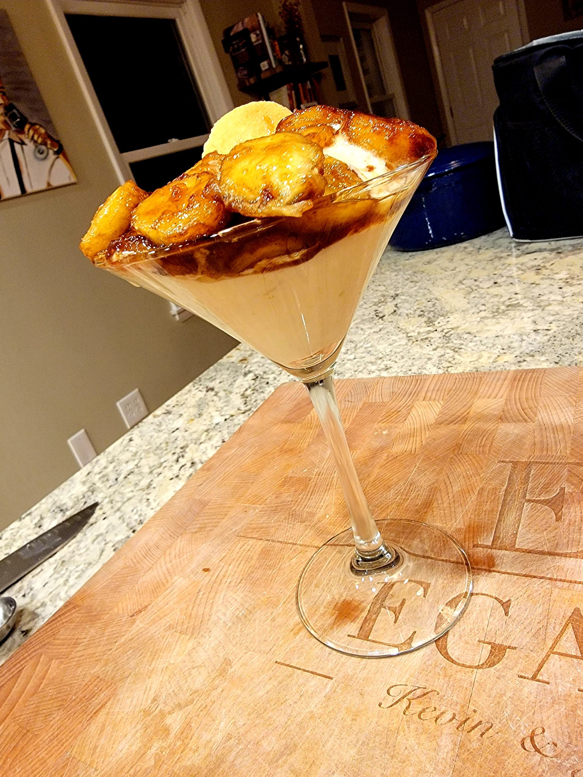 Creamy dessert in a martini glass topped with caramelized banana slices on a wooden cutting board.