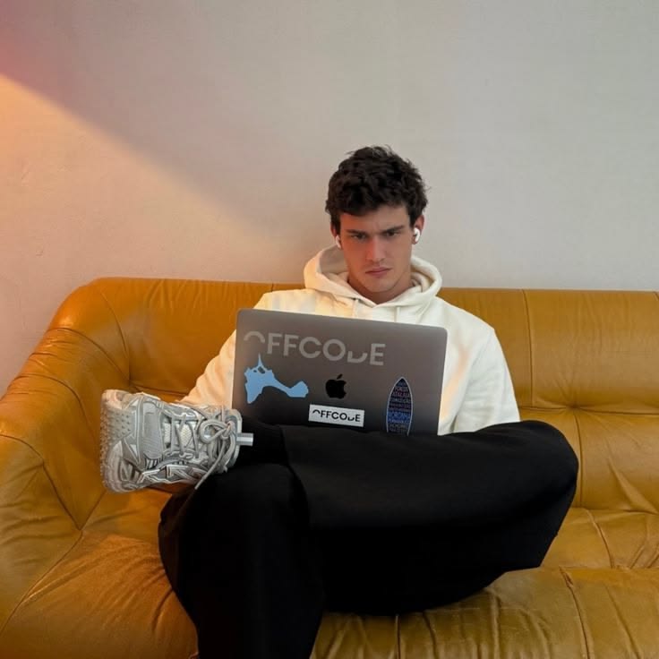 Man working on a laptop with earbuds on, on a sofa.