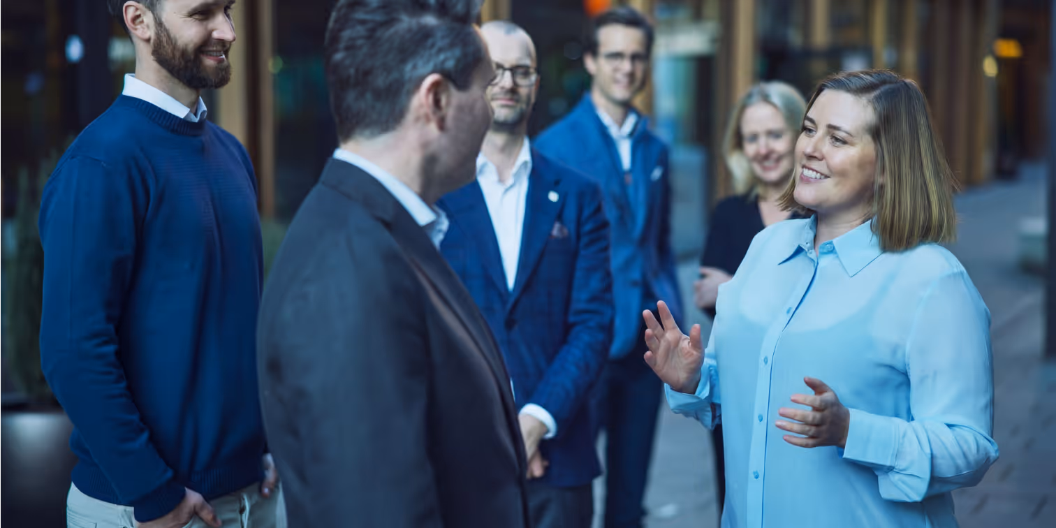 Group of six people in business casual attire having a conversation outdoors in an urban setting.