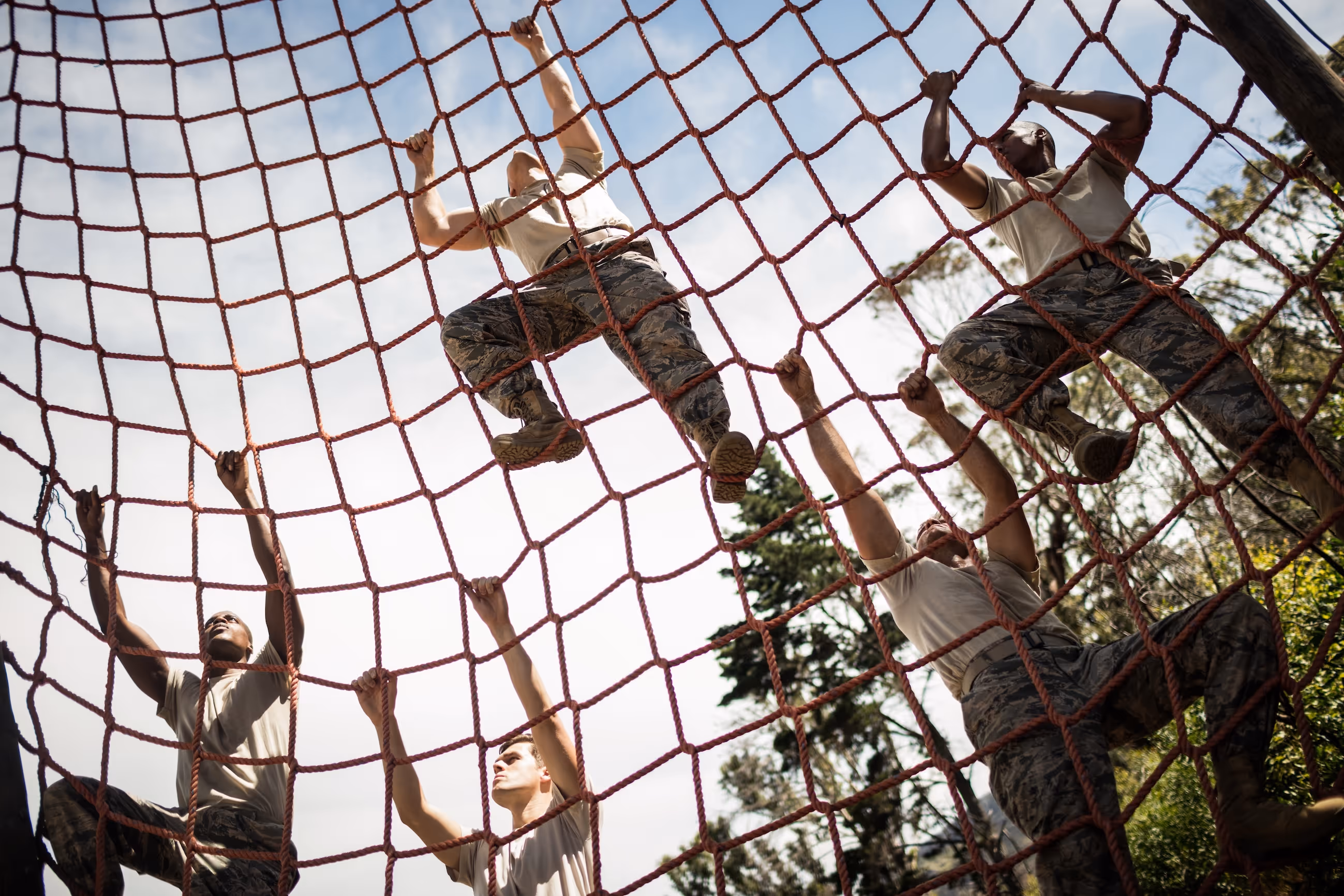 Five military personnel climbing a large red rope net outdoors during training.