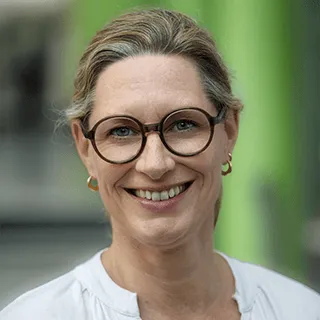 Porträt einer lächelnden Frau mit Brille und hochgestecktem Haar vor grünem Hintergrund.