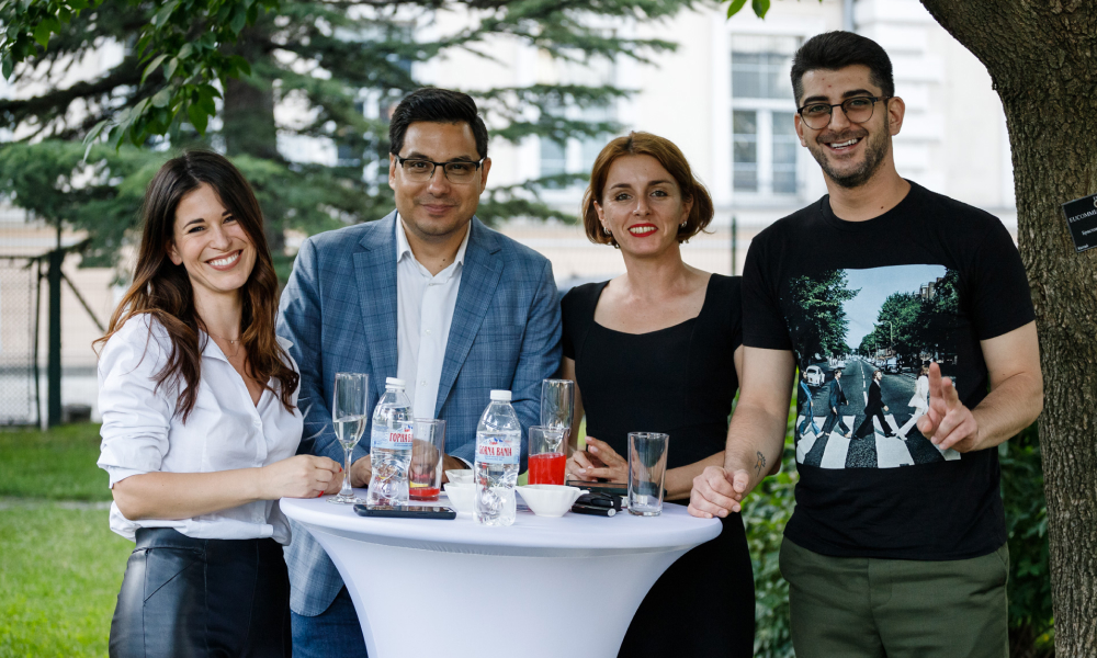 Four people smiling around a high white table outdoors with drinks and trees in the background.