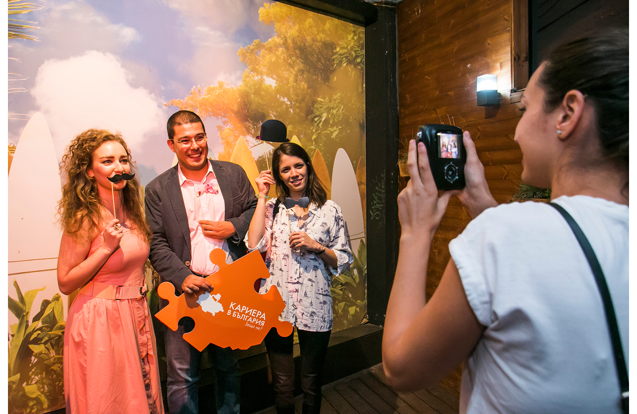 Three people pose with props for a photo taken by a fourth person indoors against a nature backdrop.