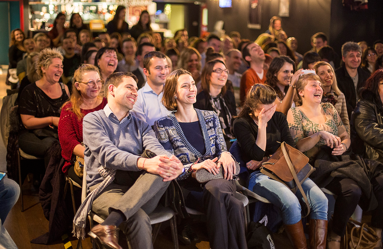 Audience sitting closely together laughing and enjoying an comedy event indoors.