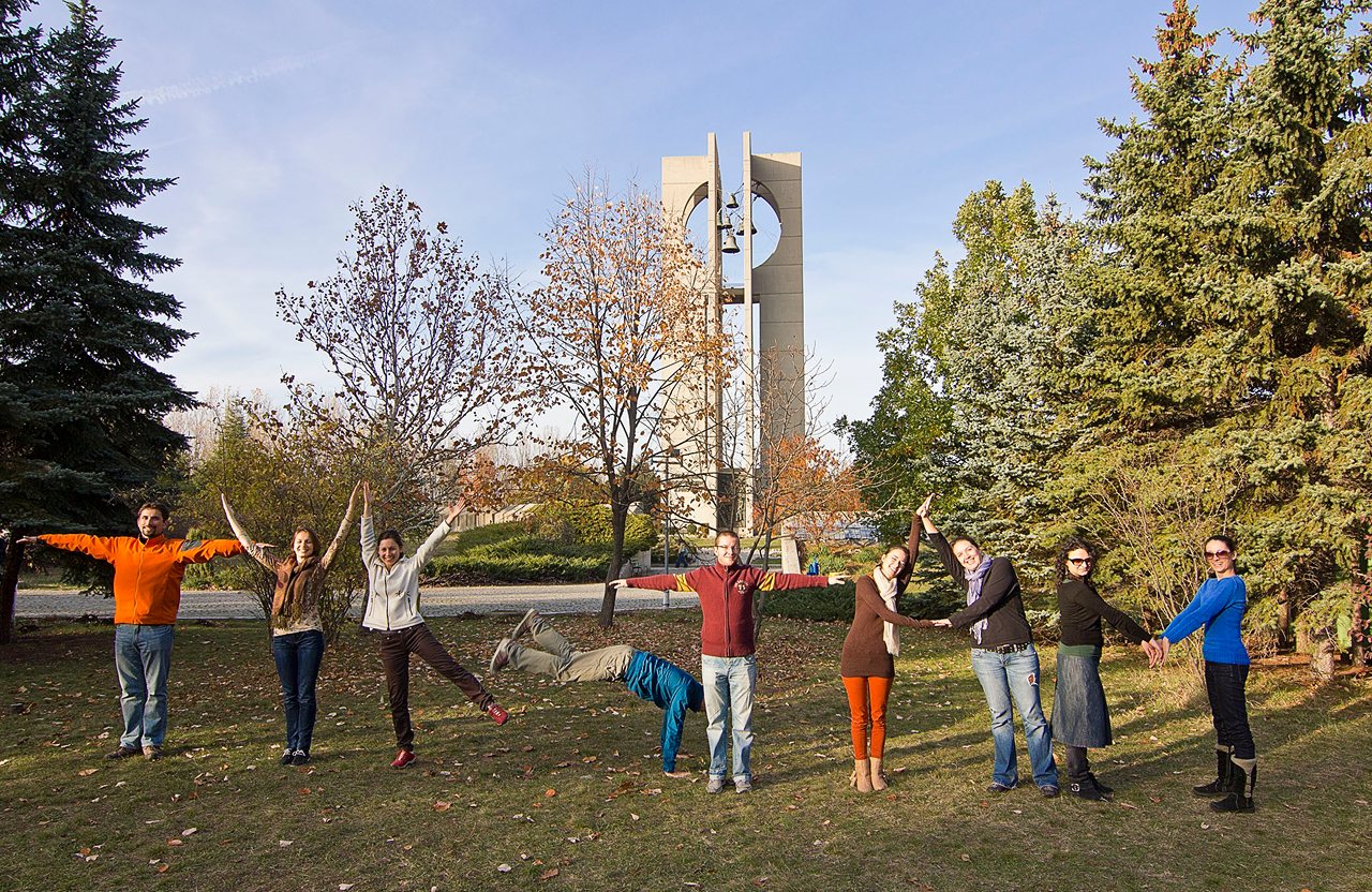 Group of nine people outdoors forming the words "Tuk-Tam" with their bodies on grass near trees and a bell tower.