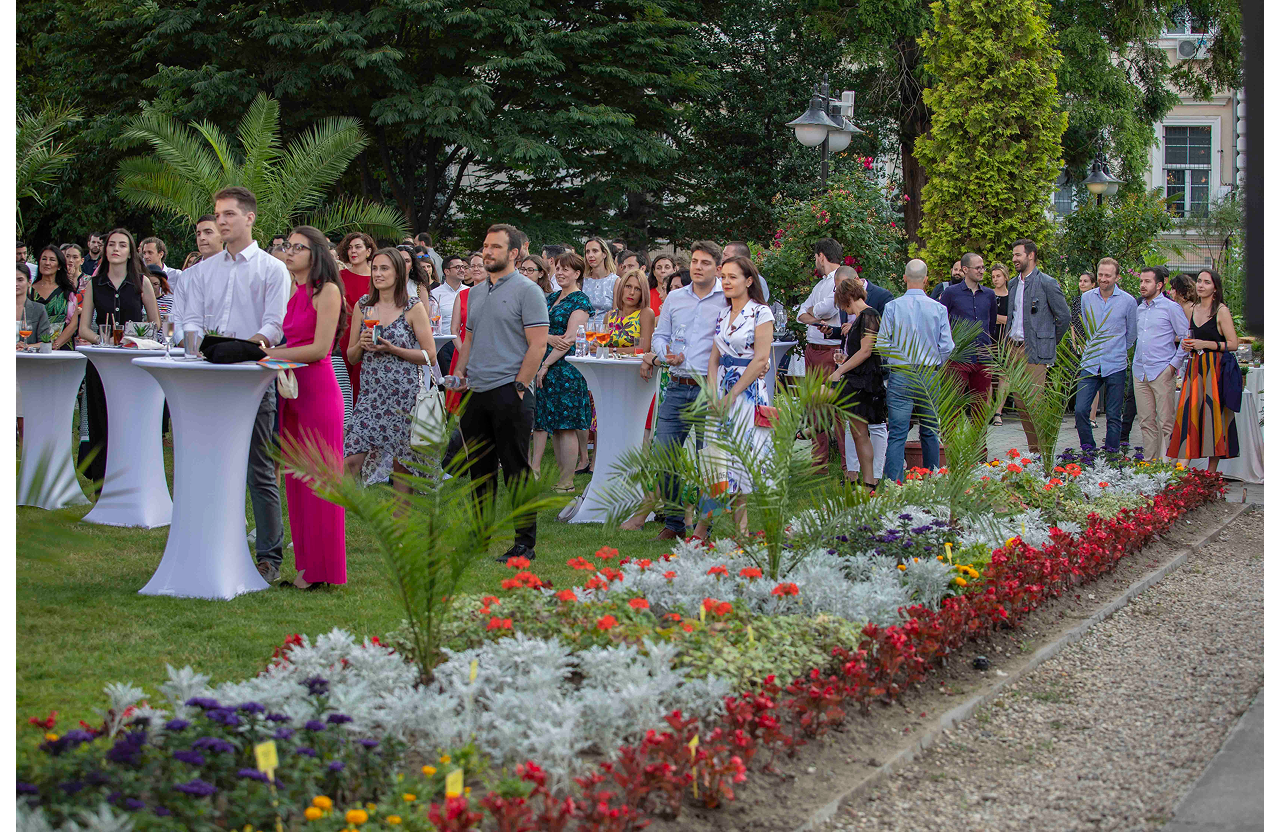 People socializing at an outdoor garden event with cocktail tables and colorful flower beds.