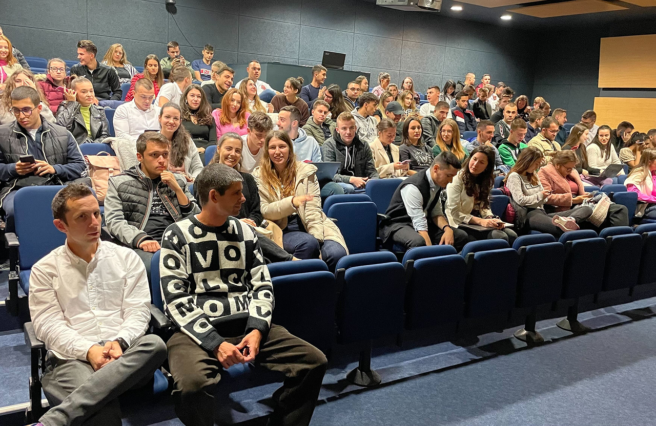 Large group of young adults seated in an auditorium, some using laptops and phones.